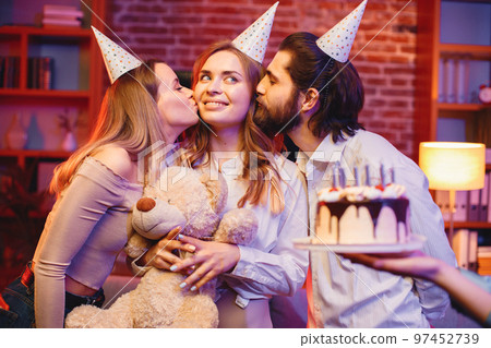 Young friends standing in indoors in evening party red light. Everybody wearing party cones. Boy and girls kissing girl in a middle in a cheek. 97452739