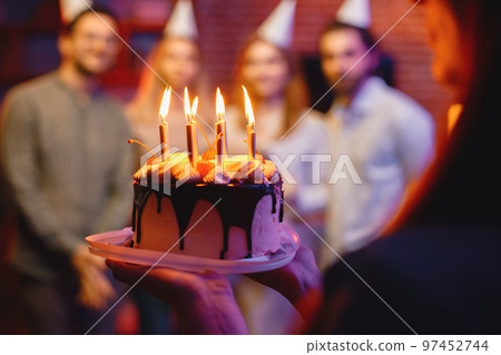 Midsection view of a woman holding a plate with birthday cake. Cake with cream, chocolates and candles. Friends in party cones standing behind blurred. Midsection view of a woman holding a plate with birthday cake. Cake with cream, chocolates and candles. Friends in party cones standing behind blurred. 97452744