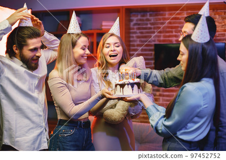 Young friends standing in indoors in evening party red light. Everybody wearing party cones. Boys and girls holding a birthday cake and light a candles. Young friends standing in indoors in evening party red light. Everybody wearing party cones. Boys and girls holding a birthday cake and light a candles. 97452752