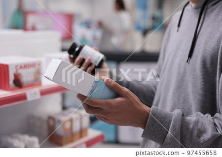 Customer looking at pills bottle and vitamins box, wanting to buy medicinal product for healthcare treatment. Person checking packages of medicaments before buying drugs. Close up. Customer looking at pills bottle and vitamins box, wanting to buy medicinal product for healthcare treatment. Person checking packages of medicaments before buying drugs. Close up. 97455658