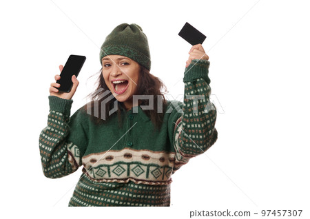 Isolated portrait on white background of a pleasant Latin American young woman, in warm woolen green clothes, smiling a toothy smile on camera, holding blank credit card and mobile phone in her hands 97457307