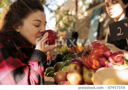 Healthy customer enjoying organic natural smell of apples, standing in front of farmers market stand. Woman smelling bio fruits before buying homegrown eco produce from counter. 97457359