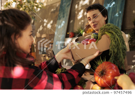 Happy smiling farmer selling healthy organic products, serving customer at farmers market. Woman buying fresh bio fruits and vegetables from local farm marketplace. Eco agriculture concept. 97457496