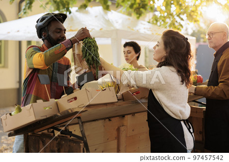 Young local vendor giving order of natural bio produce to deliveryman, helping with fruits and vegetables food delivery. Woman farmer preparing organic eco products at farmers market. Young local vendor giving order of natural bio produce to deliveryman, helping with fruits and vegetables food delivery. Woman farmer preparing organic eco products at farmers market. 97457542