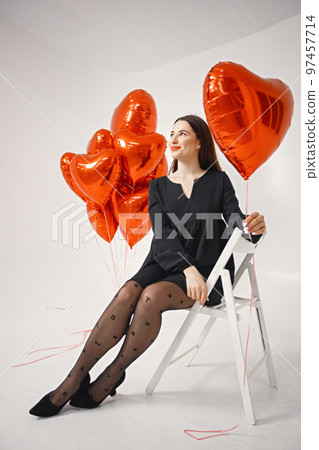 A beautiful young caucasian woman with heart shape air balloons on a white background. Woman on Valentine's Day wearing a black dress. Brunette woman in studio posing for a photo. A beautiful young caucasian woman with heart shape air balloons on a white background. Woman on Valentine's Day wearing a black dress. Brunette woman in studio posing for a photo. 97457714