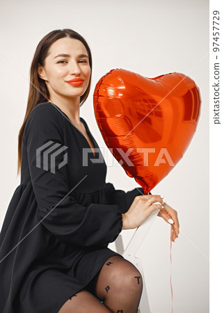 A beautiful young caucasian woman with heart shape air balloon on a white background. Woman on Valentine's Day wearing a black dress. Brunette woman in studio posing for a photo. 97457729