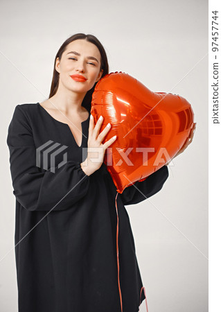 A beautiful young caucasian woman with heart shape air balloon on a white background. Woman on Valentine's Day wearing a black dress. Brunette woman in studio posing for a photo. A beautiful young caucasian woman with heart shape air balloon on a white background. Woman on Valentine's Day wearing a black dress. Brunette woman in studio posing for a photo. 97457744