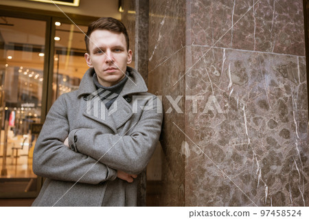 A young man stands outside a shop on the street in a gray coat leaning. A young man stands outside a shop on the street in a gray coat leaning. 97458524