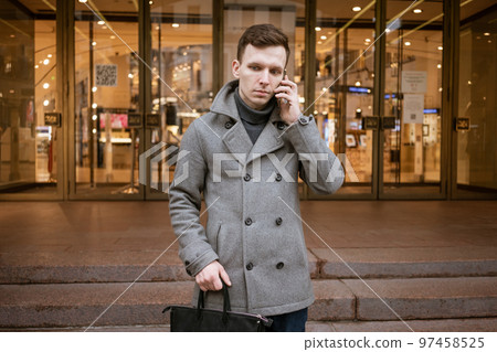 Man in gray coat and sweater stands on city street. Talks on phone. Solves 97458525