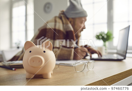 Close up of piggy bank on table with senior retired man using laptop computer in background Close up of piggy bank on table with senior retired man using laptop computer in background 97460776