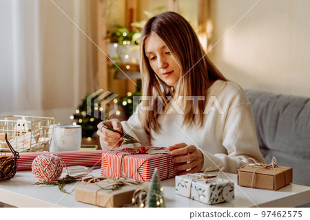 Woman wrapping Christmas eco natural gift boxes at home. Preparing presents on white table with decor elements, lights and items Christmas or New year DIY packing Concept Woman wrapping Christmas eco natural gift boxes at home. Preparing presents on white table with decor elements, lights and items Christmas or New year DIY packing Concept 97462575