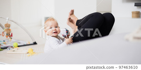 Happy family moments. Mother lying comfortably on children's mat playing with her baby boy watching and suppervising his first steps. Positive human emotions, feelings, joy 97463610