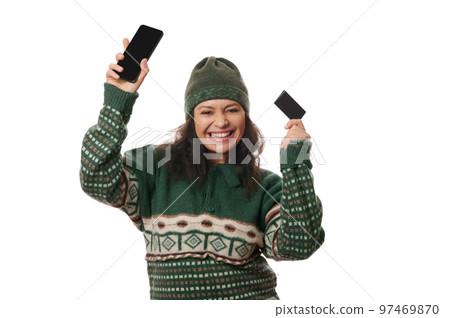 Cheeful dark-haired woman in green warm clothes posing with smartphone and credit card in her hands, smiling a beautiful toothy smile, looking at camera, isolated over white background. Copy ad space 97469870