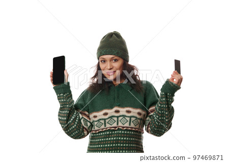 Beautiful multi-ethnic woman in warm winter wear, shows to camera a plastic bank or credit card and modern smartphone with free space for mobile apps or advertising text, isolated on white background 97469871