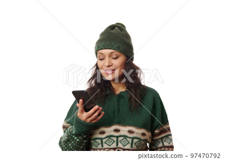 Charming multi-ethnic brunette in woolen hat and Xmas patterned green sweater, holding mobile phone and smiling cheerfully, isolated over white background with free space for your advertising text 97470792