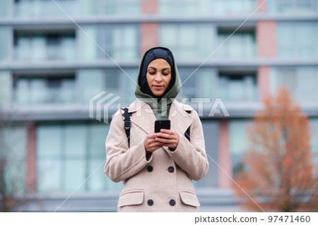 Portrait of young muslim woman in coat, standing outdoor in city, scrolling smartphone. Portrait of young muslim woman in coat, standing outdoor in city, scrolling smartphone. 97471460