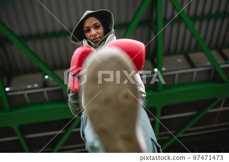 Low angle view of young muslim woman with boxing gloves standing outdoor in city. Low angle view of young muslim woman with boxing gloves standing outdoor in city. 97471473