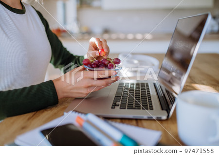 Side view of woman holding bowl with grapes above desk with computer, diary and smartphone. Work-life balance concept. Side view of woman holding bowl with grapes above desk with computer, diary and smartphone. Work-life balance concept. 97471558