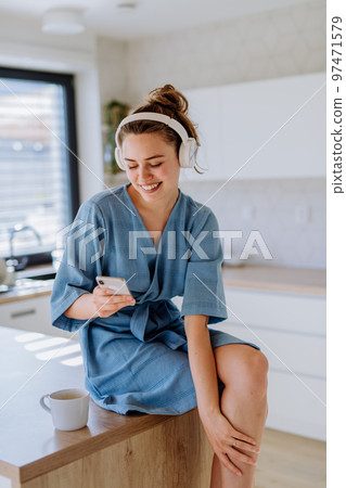 Young woman listening music, scrolling phone and enjoying cup of coffee at morning, in her kitchen. Young woman listening music, scrolling phone and enjoying cup of coffee at morning, in her kitchen. 97471579