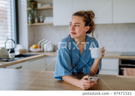 Young woman with smartphone enjoying cup of coffee at morning, in her kitchen. 97471581