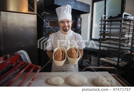 Young baker with down syndrome preparing pastries in bakery. Concept of integration people with disabilities into society. Young baker with down syndrome preparing pastries in bakery. Concept of integration people with disabilities into society. 97471787