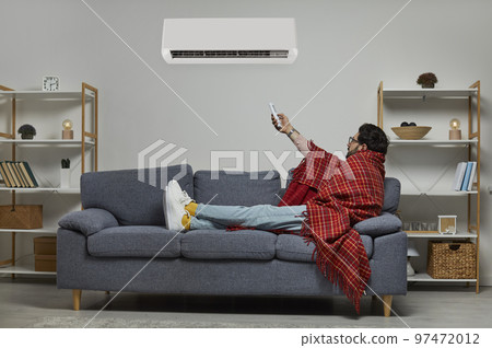 Young man wrapped in blanket holding air conditioner remote control 97472012