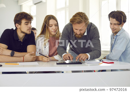 Male teacher in class helps group of highschool students working on digital tablet. 97472016