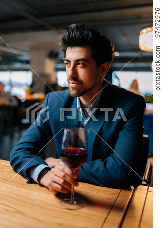 Vertical portrait of successful elegant man in fashion suit holding glasses of red wine sitting at table in restaurant, looking away. Bearded businessman resting, having dinner alone. 97472976