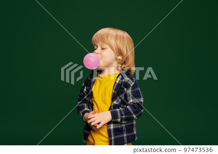 Portrait of little boy, child posing wit bubblegum over green studio background. Having fun. Concept of childhood, emotions 97473536