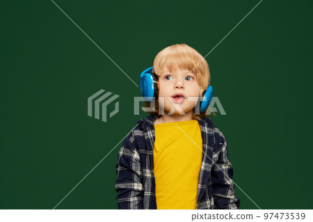 Portrait of little boy, child posing in headphones, listening to fairy tales over green studio background. Concept of childhood, emotions 97473539