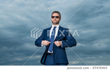 Businessman wearing sunglasses sky background. Grizzled businessman buttoning suit jacket Businessman wearing sunglasses sky background. Grizzled businessman buttoning suit jacket 97476403