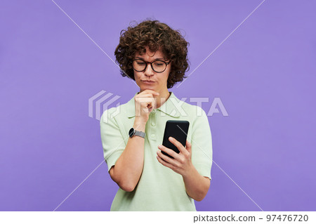 Young pensive woman with smartphone looking through goods in online shop 97476720