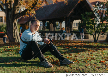 Professional Photographer Taking Picture Of Beautiful Autumn Park. Man Professional Photographer Sit With Camera And With Smartphone In Autumn Park. Retouched, Vibrant Colors, Brownish Tones. Professional Photographer Taking Picture Of Beautiful Autumn Park. Man Professional Photographer Sit With Camera And With Smartphone In Autumn Park. Retouched, Vibrant Colors, Brownish Tones. 97477243