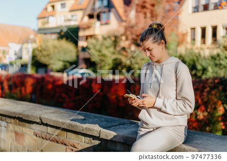 Young Fashionable Teenage Girl With Smartphone In Europian Park In Autumn Sitting At Smiling. Trendy Young Woman In Fall In Park Texting. Retouched, Vibrant Colors. Beautiful Blonde Teenage Girl Young Fashionable Teenage Girl With Smartphone In Europian Park In Autumn Sitting At Smiling. Trendy Young Woman In Fall In Park Texting. Retouched, Vibrant Colors. Beautiful Blonde Teenage Girl 97477336