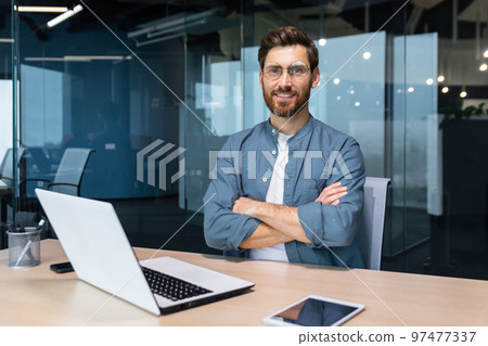 Portrait of mature businessman with beard, man in shirt smiling and looking at camera sitting at desk with crossed arms inside modern office building, using tablet and laptop at work. Portrait of mature businessman with beard, man in shirt smiling and looking at camera sitting at desk with crossed arms inside modern office building, using tablet and laptop at work. 97477337