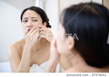 Problem skin. Concerned young asian women popping pimple on cheek while standing near mirror in bathroom. young asian women with acne Problem skin. Concerned young asian women popping pimple on cheek while standing near mirror in bathroom. young asian women with acne 97478042