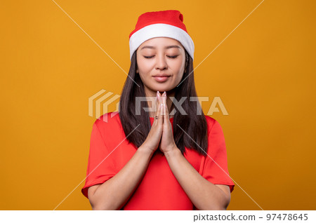 Young woman in santa hat praying with her eyes closed Young woman in santa hat praying with her eyes closed 97478645