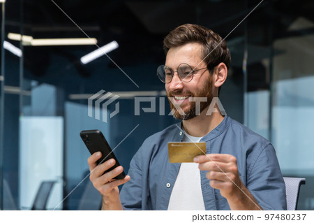 Cheerful and smiling bearded businessman doing online shopping and bank money transfer, freelancer holding bank credit card and smartphone, man sitting at desk inside office. Cheerful and smiling bearded businessman doing online shopping and bank money transfer, freelancer holding bank credit card and smartphone, man sitting at desk inside office. 97480237