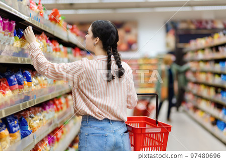 Back view of caucasian woman choosing products in grocery store. Shelves with food in background. Concept of shopping and consumerism Back view of caucasian woman choosing products in grocery store. Shelves with food in background. Concept of shopping and consumerism 97480696