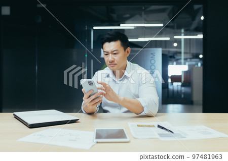 Worried young man Asian businessman sits in the office at the table, holds the phone in his hands, dials a message, is upset and reads bad news. Worried young man Asian businessman sits in the office at the table, holds the phone in his hands, dials a message, is upset and reads bad news. 97481963