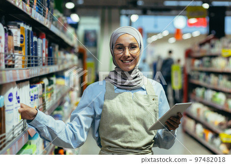 Portrait of a female saleswoman in a hijab, a salesperson in a household chemicals department is smiling and looking at the camera, holding a laptop tablet computer in her hands. Portrait of a female saleswoman in a hijab, a salesperson in a household chemicals department is smiling and looking at the camera, holding a laptop tablet computer in her hands. 97482018