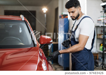 Bearded young male inspector measuring paint cover thickness of the car body at the vehicle service Bearded young male inspector measuring paint cover thickness of the car body at the vehicle service 97483426