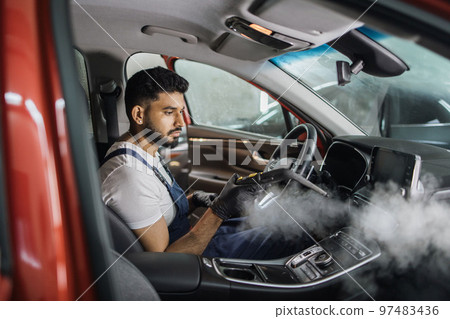 Handsome bearded man in blue uniform overalls cleaning interior of the car with hot steam cleaner. 97483436
