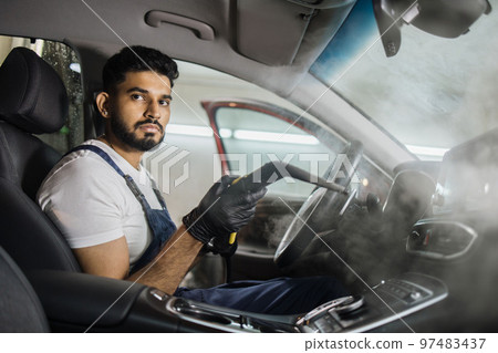 Handsome man in overalls, worker of car wash center, cleaning car interior with hot steam cleaner. 97483437