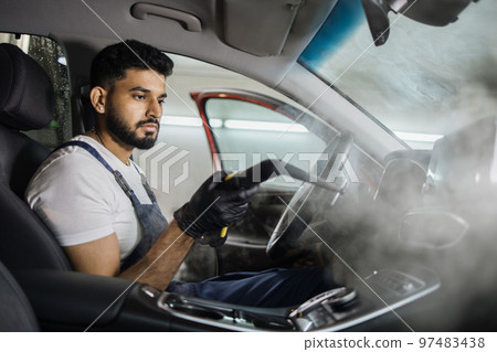 Handsome man in overalls, worker of car wash center, cleaning car interior with hot steam cleaner. 97483438