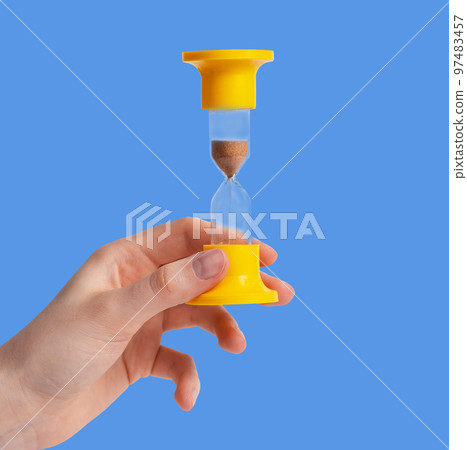 Yellow hourglass with running sand in female hand over blue background 97483457