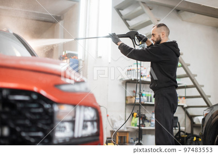 Portrait of of young bearded man worker washing red car under high pressure water Portrait of of young bearded man worker washing red car under high pressure water 97483553