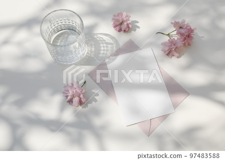 Wedding composition. Feminine spring breakfast still life. Pink Japanese cherry tree, sakura blossoms in sunlight. Blank greeting card, invitation. Glass of water. White dappled table background. 97483588