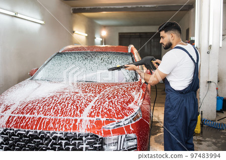 Handsome young bearded man worker, wearing protective clothes and gloves, washing modern red car 97483994