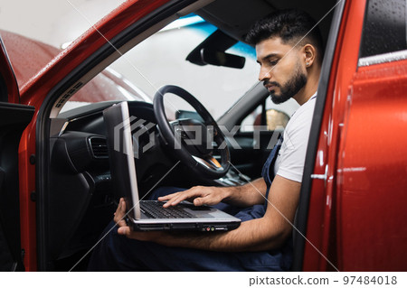 Bearded male mechanic sitting inside car using laptop, recording automobile engine checks Bearded male mechanic sitting inside car using laptop, recording automobile engine checks 97484018